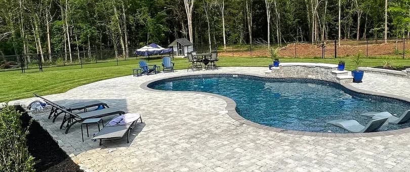 A finished freeform backyard pool with a paver deck, retaining wall, and fire pit, backing onto a Middle Tennessee treeline.